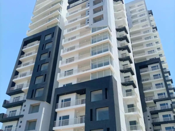 Modern High-Rise Apartment Tower With Black And White Facade, Glass Balconies, Large Windows, And Landscaped Base Under Blue