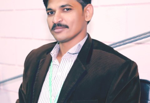 Portrait Of A Man In A Dark Blazer And Striped Shirt Seated Against A Light Interior Background
