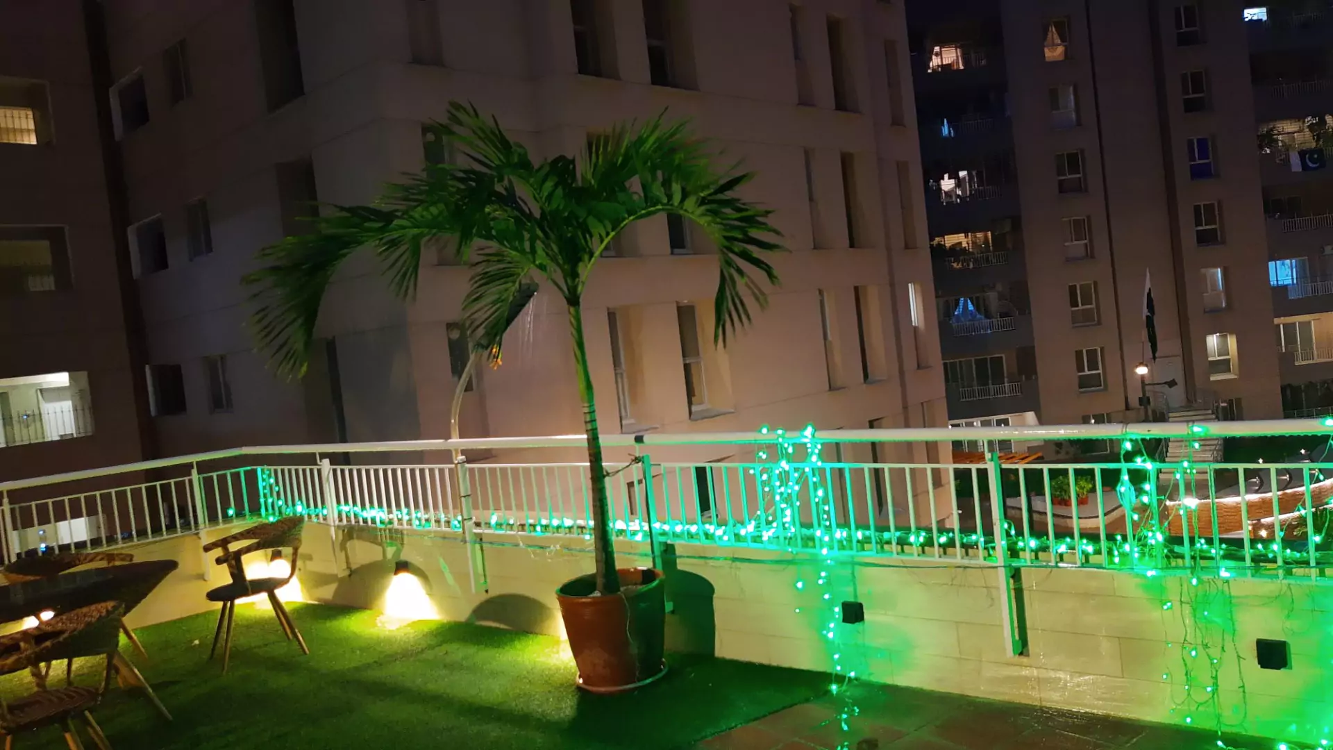 Luxury Flats & Penthouses For Sale in Lakhani Presidency (4) Lakhani Presidency balcony in Karachi with potted palm tree, seating area, artificial turf, green LED lights, and high-rise v