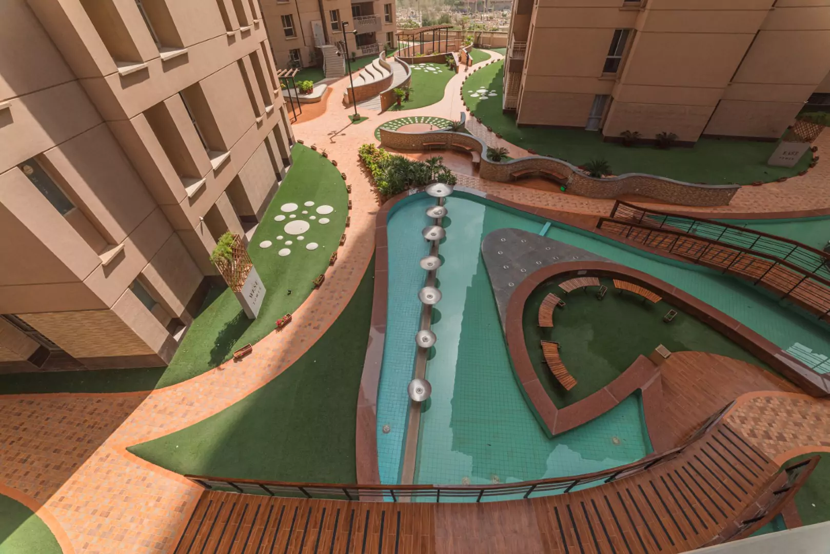 Luxury Flats & Penthouses For Sale in Lakhani Presidency (6) Elevated view of Lakhani Presidency courtyard in Karachi with curved pool, wooden walkways, green lawns, pebble paths, and be