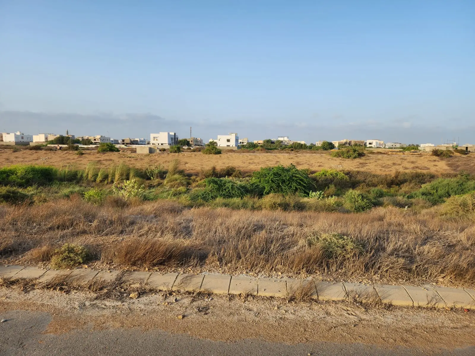 Prime Commercial Plot for Sale in Peninsula Commercial, DHA Phase 8 Karachi (1) Vacant commercial plot in Peninsula Commercial DHA Phase 8 with sandy terrain, dry bushes, concrete curb, and distant white b