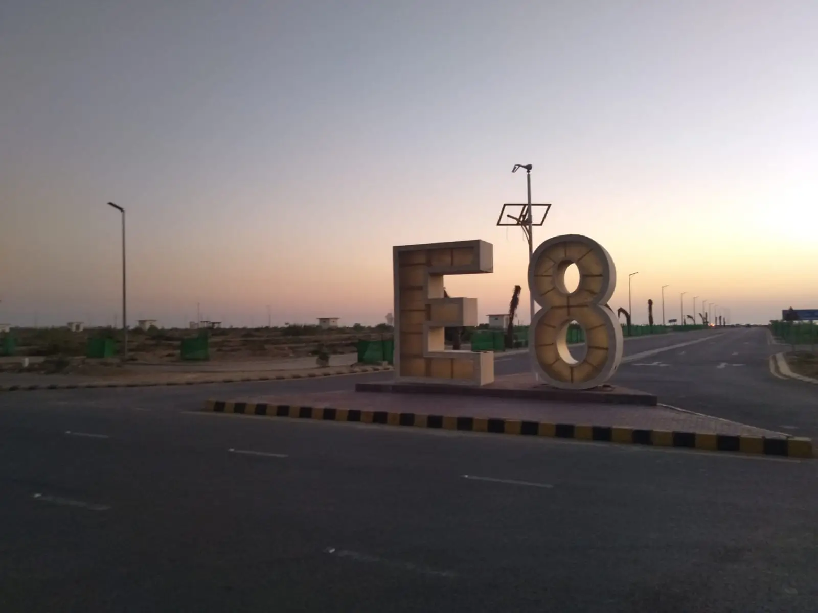 Prime Commercial Plot for Sale in Peninsula Commercial, DHA Phase 8 Karachi (3) E8 sign at Peninsula Commercial DHA Phase 8 entrance with wide roads, streetlights, barriers, and sunset sky