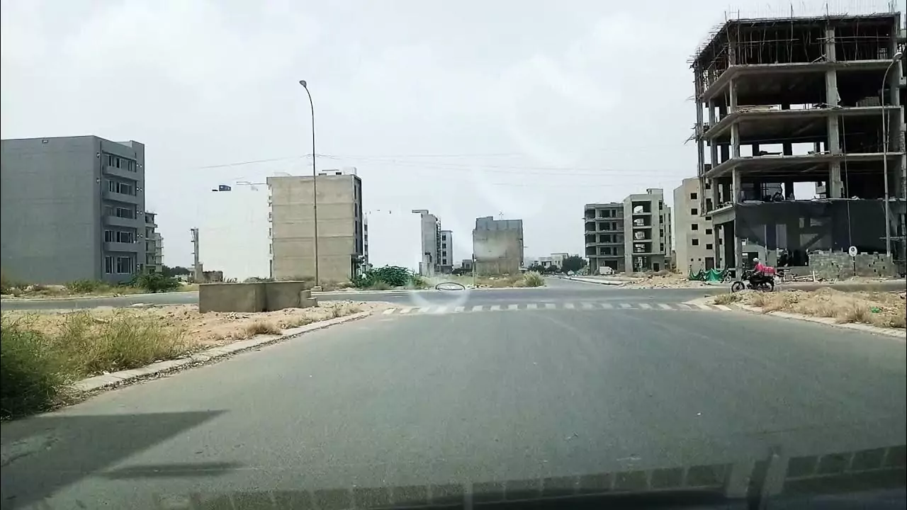 Prime Commercial Plot for Sale in Peninsula Commercial, DHA Phase 8 Karachi Wide road intersection in Peninsula Commercial DHA Phase 8 with under-construction concrete buildings, streetlights, and pede