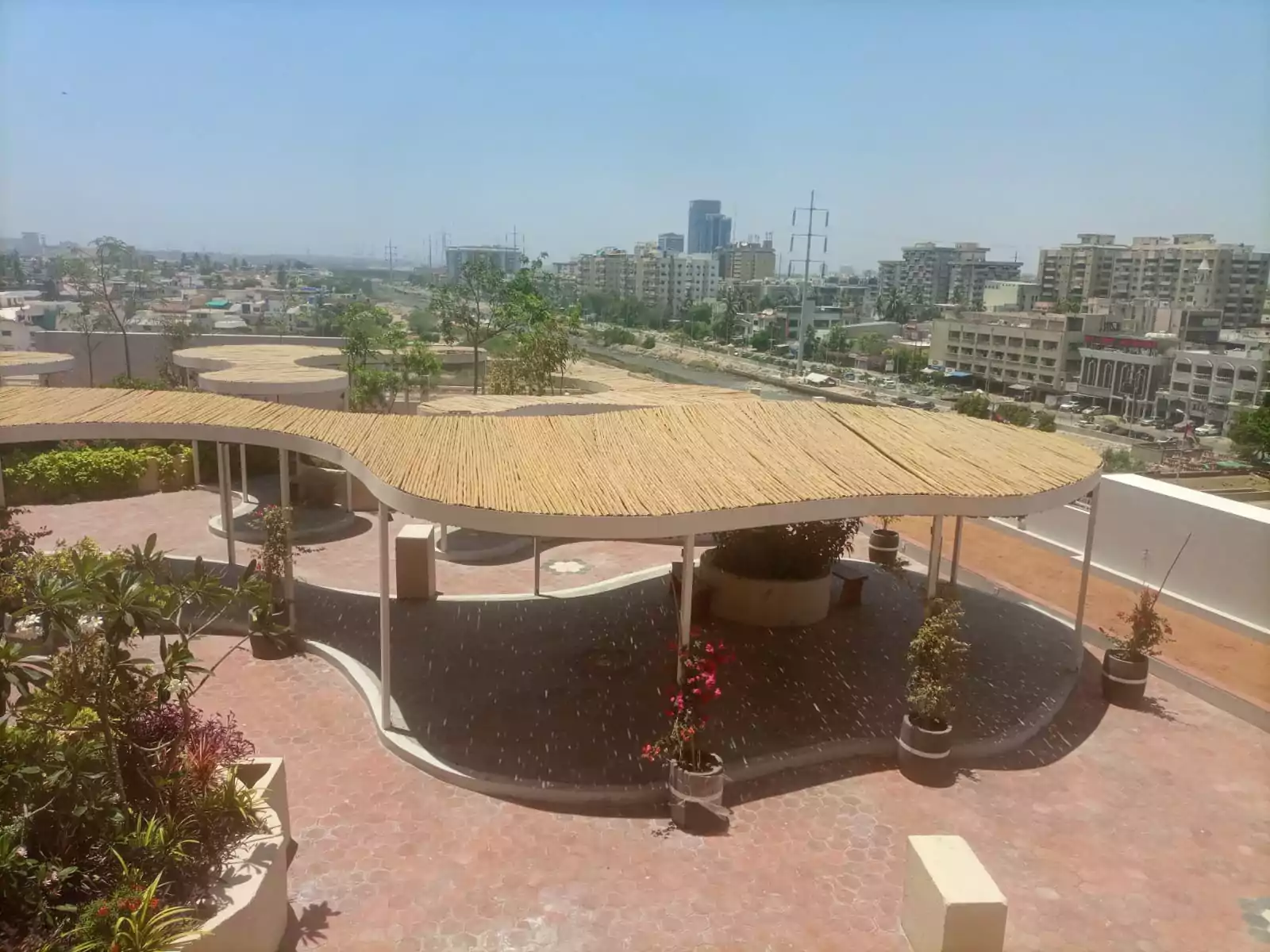 Dynasty Apartments Clifton Luxury 4-Bedroom Flats for Sale in Karachi (2) Rooftop terrace with wavy thatched pergola, potted plants and greenery at Dynasty Apartments Clifton, city skyline view