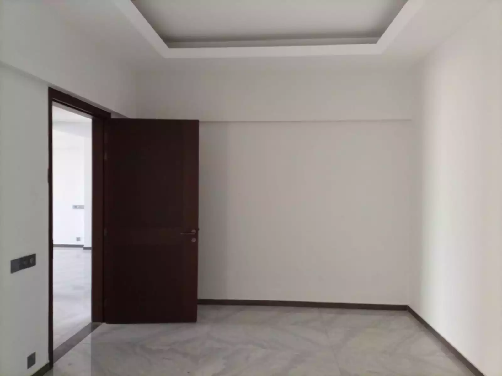 Dynasty Apartments Clifton Luxury 4-Bedroom Flats for Sale in Karachi (3) Spacious empty bedroom in Dynasty Apartments Clifton with white walls, false ceiling, brown wooden door, and gray marble floo