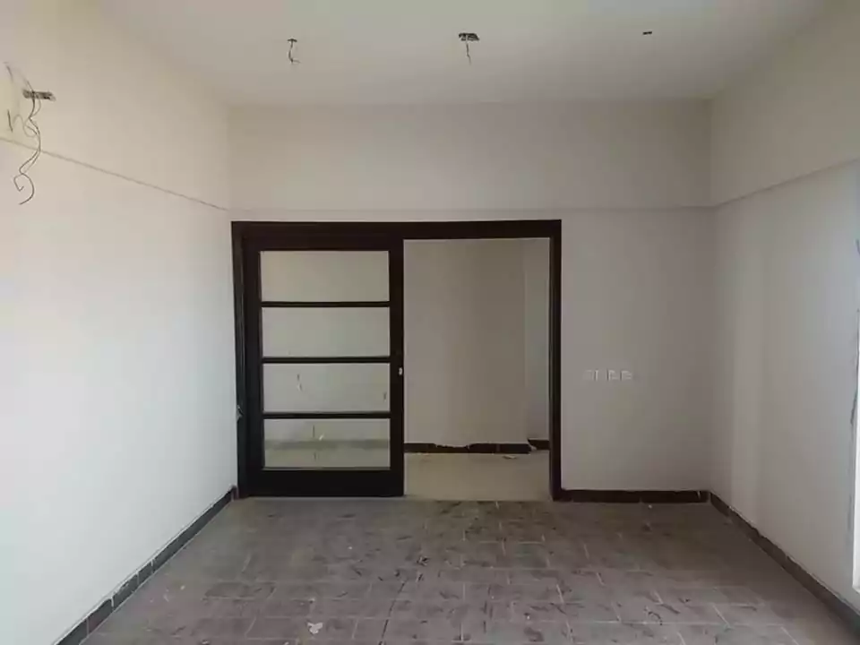 Luxurious 3-Bedroom Apartment for Sale in Green One Clifton Block 8 Karachi (5) Spacious empty room in Greens One Clifton 3-bedroom apartment with gray tiled floor, white walls, and black-framed sliding gl