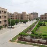 Aerial View Of Multi-Story Beige Apartment Buildings, Central Paved Road, Lush Green Lawns, Trees, And Park In Residential Co