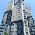 Modern High-Rise Apartment Tower With Black And White Facade, Glass Balconies, Large Windows, And Landscaped Base Under Blue