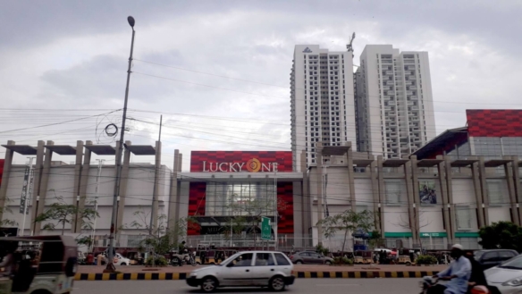Lucky One Apartments Exterior With Tall White High-Rise Towers, Red Lucky One Mall Facade, Street Lamps, Cars, And Pedestrian