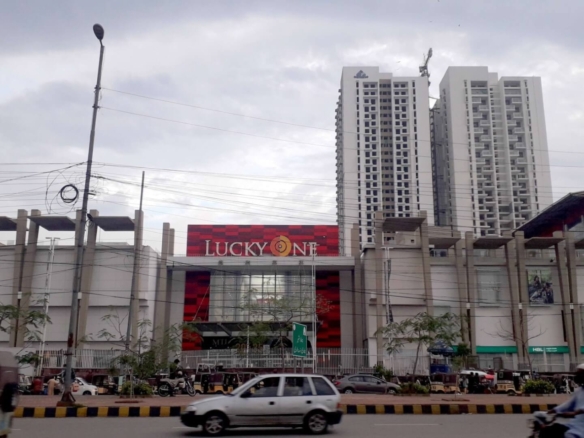 Lucky One Apartments Exterior With Tall White High-Rise Towers, Red Lucky One Mall Facade, Street Lamps, Cars, And Pedestrian