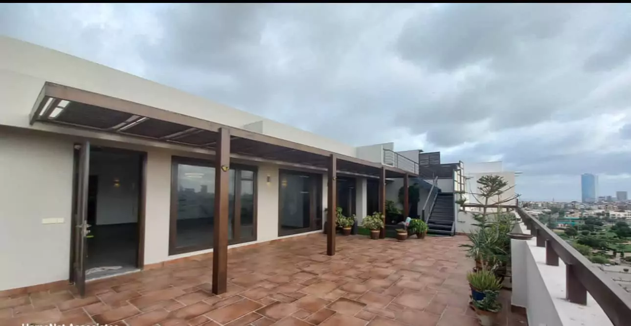 Luxurious 5-Bed Penthouse for Rent in Mall Square Zamzam – DHA Phase 5 Karachi (12) Spacious terrace with wooden pergola, glass doors, potted plants, tiled flooring and city skyline view in 5-bed penthouse, Ma
