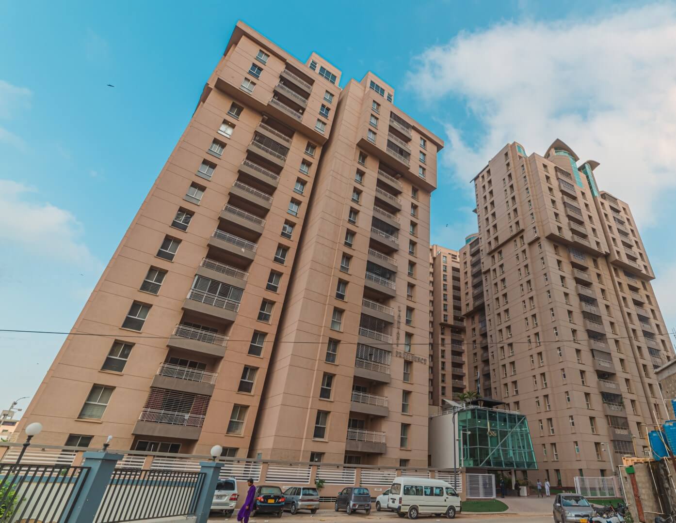 Tall modern beige apartment towers with balconies at Lakhani Presidency in Karachi, gated entrance and parked vehicles