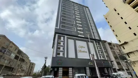 Modern Multi-Story Clifton Mansion Building In Karachi With Grey Facade, Balconies, Prominent Signage, Street Entrance And Pa