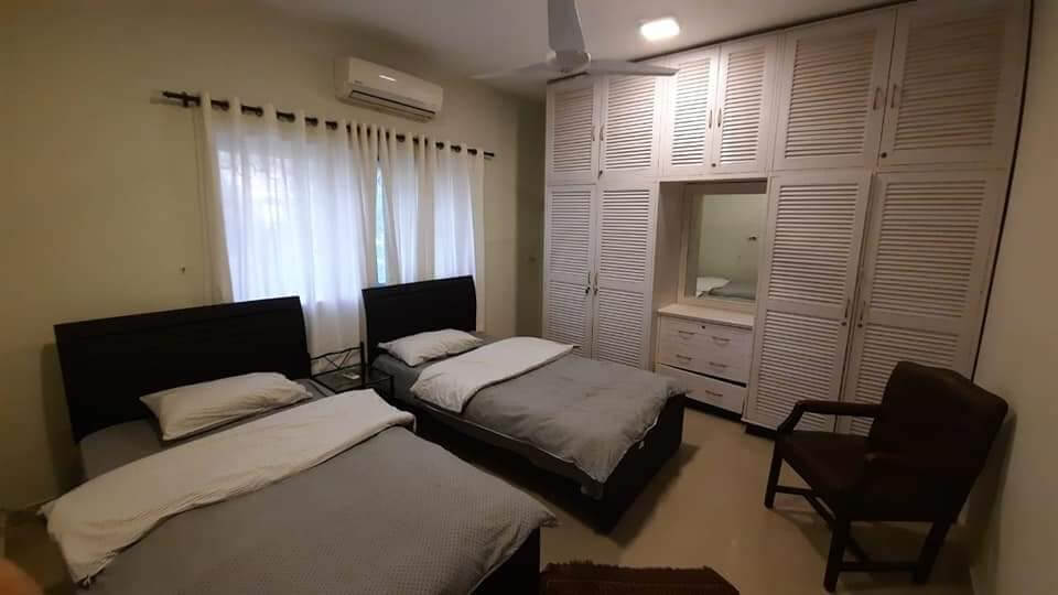 Fully Furnished Portion for Rent in DHA Phase 4, Karachi (4) Twin bedroom with two single beds, large louvered wardrobe, AC unit, ceiling fan, windows with sheer curtains, and armchair i