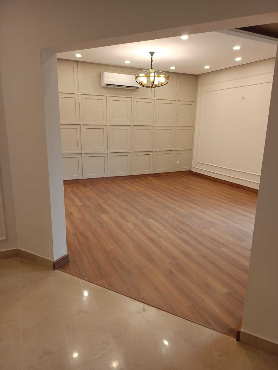 Brand New Stylish Bungalow For Rent in DHA Phase 4 Karachi (2) Spacious empty lounge with wooden flooring, paneled beige walls, chandelier, recessed lights, AC unit in brand new bungalow f