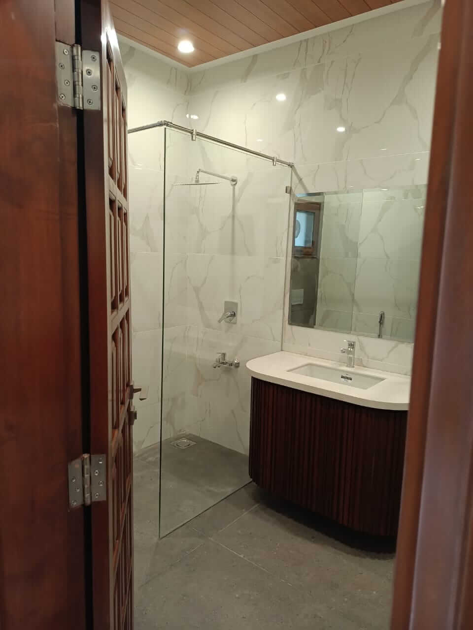 Brand New Stylish Bungalow For Rent in DHA Phase 4 Karachi (9) Modern bathroom with glass shower enclosure, white marble tiles, curved wooden vanity sink, open wooden door in brand new sty
