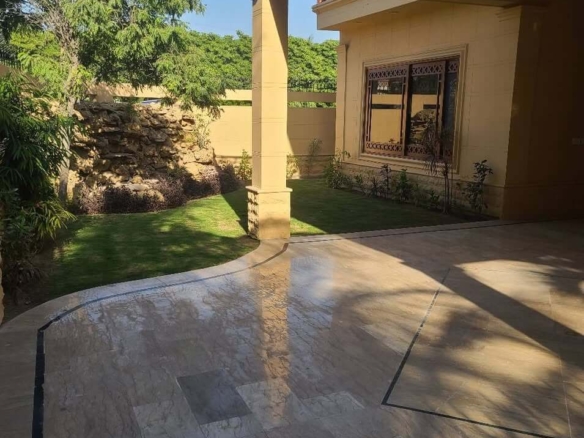 Luxury Bungalow Exterior In Dha Phase 6 With Beige Tiled Patio, Green Lawn, Rock Garden, Pillars, And Arched Windows