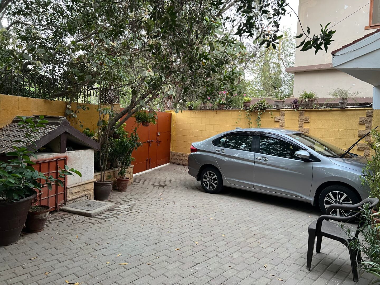 Portion for Rent in DHA Karachi – Fully Furnished Prime Location MaxX Capitals (14) Paved parking area of portion for rent in DHA Karachi with parked silver sedan, yellow walls, potted plants, tree, wooden doo
